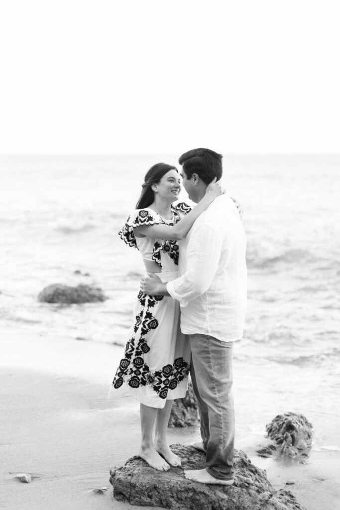 El Matador Beach Engagement Session - A couple stands barefoot on rocks at the beach, smiling and looking at each other. The woman wears a dress with a floral pattern and the man wears a white shirt and pants. Ocean waves and sand are visible in the background. Copyright - Lisa Leanne Photography, California