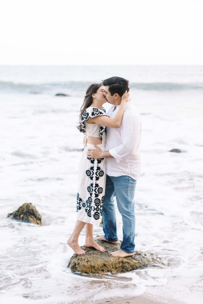 El Matador Beach Engagement Session - A couple stands barefoot on a rock at the beach, embracing and kissing with waves gently surrounding them. The woman wears a white dress with black patterns, and the man wears a white shirt and jeans. Copyright - Lisa Leanne Photography, California