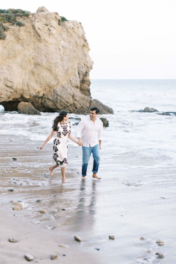 El Matador Beach Engagement Session - A couple walks barefoot along a sandy beach, holding hands. The woman wears a white dress with black floral patterns, and the man wears a white shirt and jeans. Waves wash up on shore, and a large rocky cliff is in the background. Copyright - Lisa Leanne Photography, California