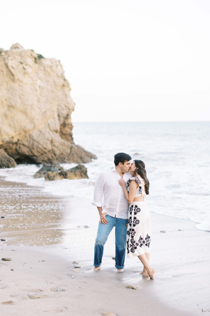 El Matador Beach Engagement Session - A couple kisses on a sandy beach near the ocean, with rocky cliffs in the background. The man wears a white shirt and jeans, and the woman wears a white dress with black patterns. Copyright - Lisa Leanne Photography, California
