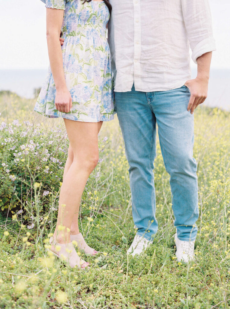 Malibu Beach Engagement Session - A couple stands together outdoors in a field of grass and wildflowers. The woman wears a short floral dress and the man wears a white shirt and blue jeans. Their faces are not visible. Copyright - Lisa Leanne Photography, California