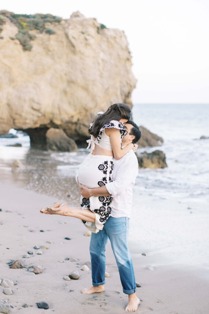 El Matador Beach Engagement Session - A man in jeans lifts and embraces a woman in a patterned dress on a rocky beach. They are smiling, barefoot, and facing each other, with waves and a large cliff in the background. Copyright - Lisa Leanne Photography, California