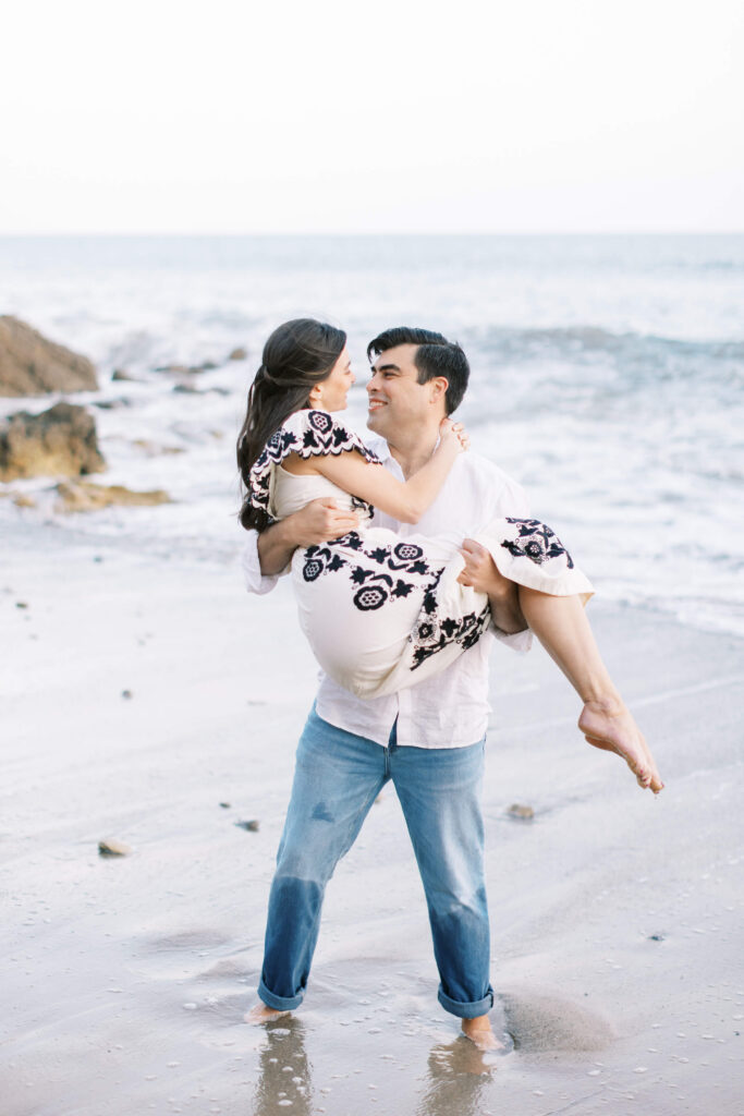 El Matador Beach Engagement Session - A man wearing jeans and a white shirt stands on a beach, smiling as he carries a woman in a patterned dress. The woman looks at him and smiles, with the ocean and rocks in the background. Copyright - Lisa Leanne Photography, California