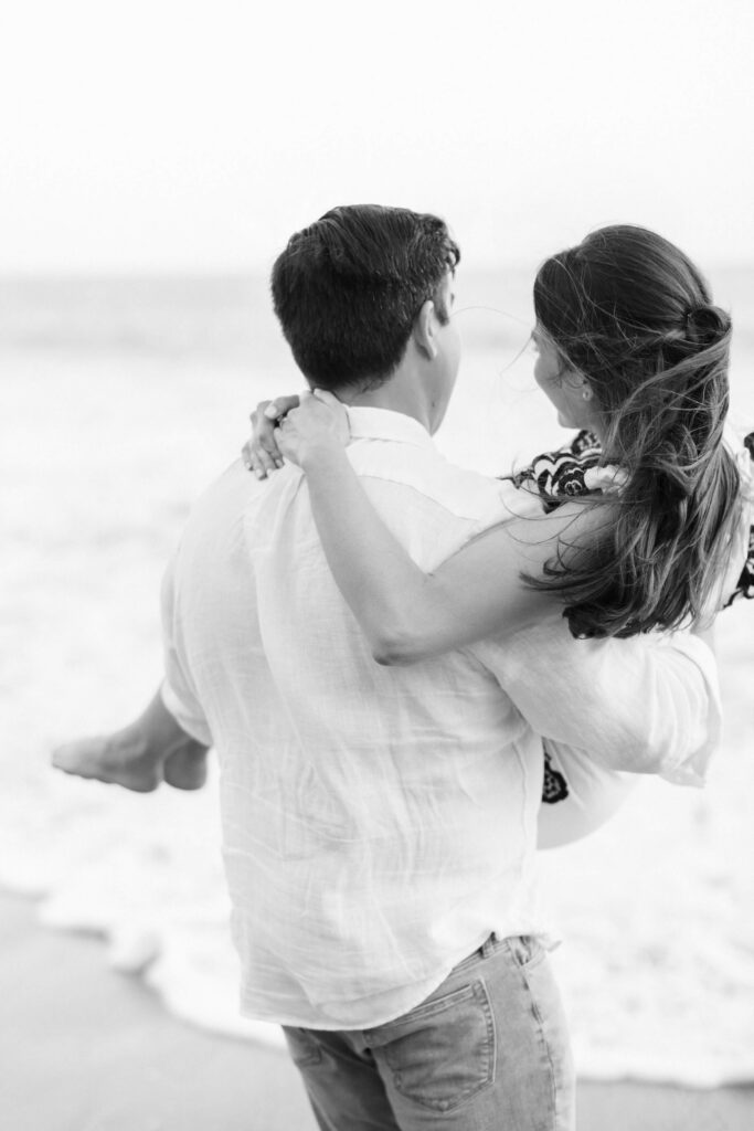 El Matador Beach Engagement Session - A man stands on the beach, holding a woman in his arms. They both face the ocean, smiling, with the woman’s hair blowing in the breeze. The image is in black and white. Copyright - Lisa Leanne Photography, California