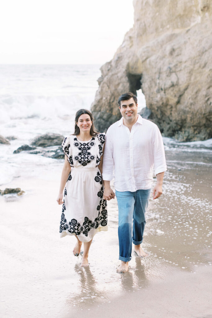 El Matador Beach Engagement Session - A smiling couple walks barefoot on the beach near rocky cliffs. The woman wears a white dress with black floral patterns, and the man wears a white shirt with jeans. Ocean waves and wet sand are visible around them. Copyright - Lisa Leanne Photography, California