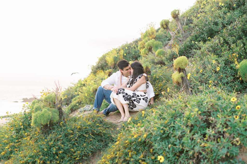 Springtime El Matador Beach Engagement Session - A couple sits closely together on a grassy hillside covered in green plants and yellow flowers, gazing at each other affectionately. The scene is peaceful and sunlit, with the ocean visible in the background. Copyright - Lisa Leanne Photography, California