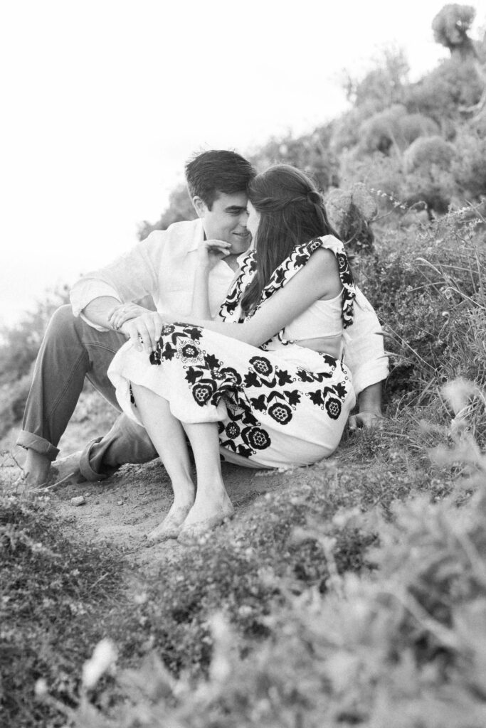 Springtime El Matador Beach Engagement Session - A black-and-white photo of a couple sitting closely together on a grassy slope, smiling and gazing at each other affectionately. The woman wears a patterned dress, and the man wears rolled-up pants and a light shirt. Copyright - Lisa Leanne Photography, California