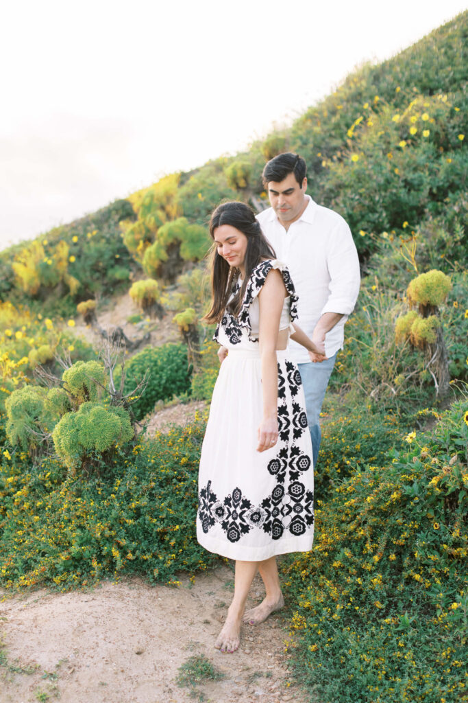 Springtime El Matador Beach Engagement Session - A woman in a white dress with black floral patterns walks barefoot on a dirt path, followed by a man in a white shirt and jeans, surrounded by green plants and yellow flowers on a hillside. Copyright - Lisa Leanne Photography, California