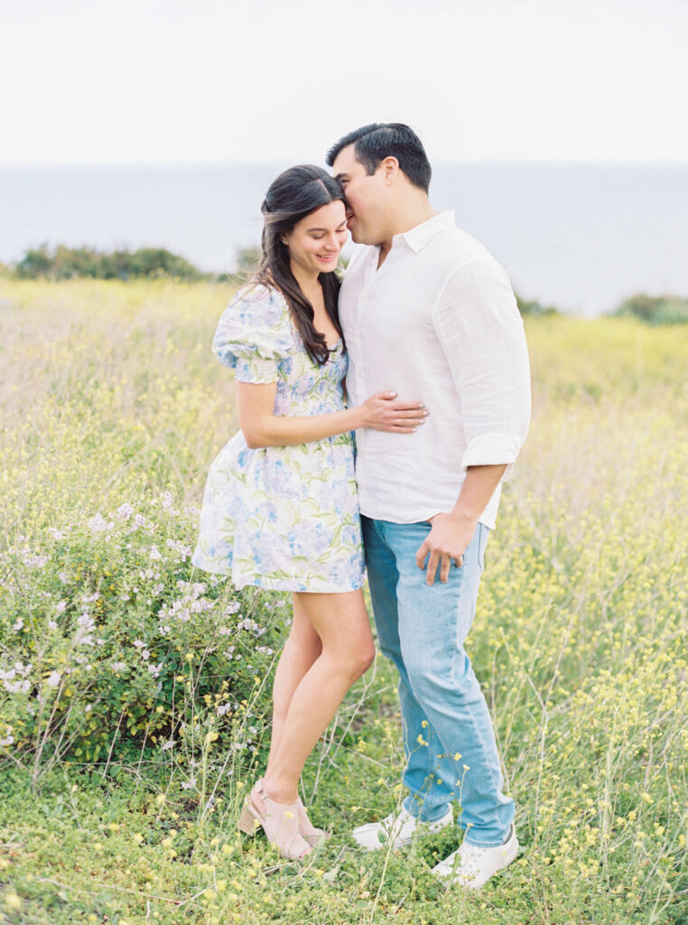 Springtime Malibu Beach Engagement Session - A couple stands together in a sunny, grassy field near the ocean. The woman in a floral dress smiles while the man in a white shirt and jeans leans in to kiss her forehead. Both appear happy and relaxed. Copyright - Lisa Leanne Photography, California