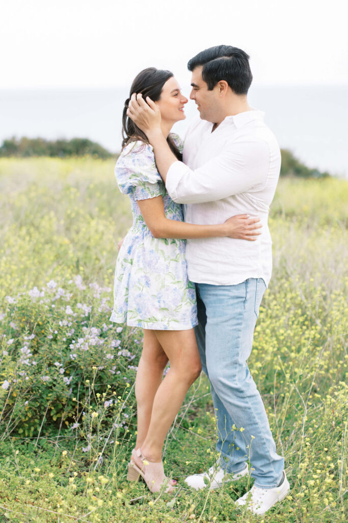 Springtime Malibu Beach Engagement Session - A couple stands close together in a grassy field, smiling and looking into each other's eyes. The woman wears a floral dress and the man is in a white shirt and jeans. Wildflowers and greenery surround them. Copyright - Lisa Leanne Photography, California