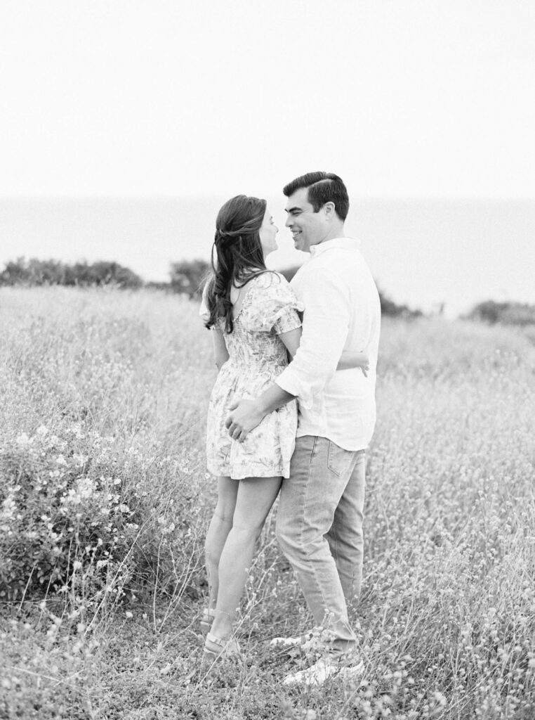 Springtime Malibu Beach Engagement Session - A couple stands together in a grassy field, facing each other and smiling. The woman wears a floral dress and the man wears a light shirt and jeans. The background shows wildflowers and a blurred view of the ocean. Copyright - Lisa Leanne Photography, California