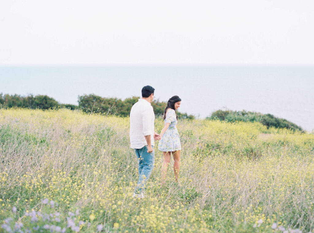 Malibu Beach Engagement Session - A couple walks hand in hand through a grassy field with yellow wildflowers, with the ocean and a clear sky visible in the background. Copyright - Lisa Leanne Photography, California