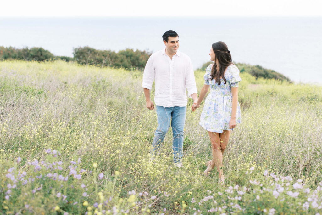Malibu Beach Engagement Session - A couple holding hands walks through a grassy field with wildflowers, smiling at each other. The ocean and shrubs are visible in the background under a pale sky. Copyright - Lisa Leanne Photography, California