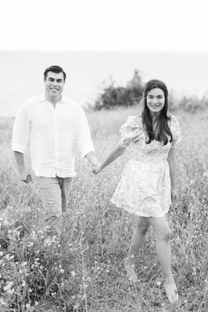 Malibu Beach Engagement Session - A couple holding hands walks through a grassy field with wildflowers. Both are smiling; the man wears a light shirt and pants, and the woman wears a floral dress. The background shows blurred trees and the horizon. Copyright - Lisa Leanne Photography, California