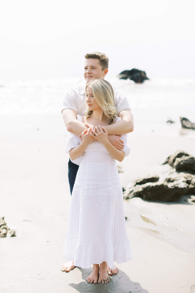 Malibu Engagement Session -A couple stands barefoot on a sandy beach. The man embraces the woman from behind, both wearing white clothes. They face different directions, with rocks and the ocean in the background under bright daylight. Copyright - Lisa Leanne Photography, California
