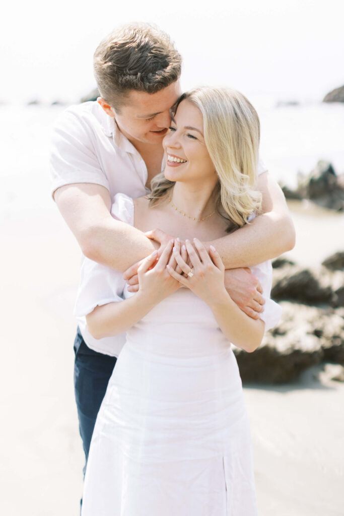 Malibu Engagement Session - A man embraces a smiling woman from behind on a sandy beach, both dressed in white. The woman rests her hands on his arms, and they appear happy, with the ocean and rocks in the background. Copyright - Lisa Leanne Photography, California