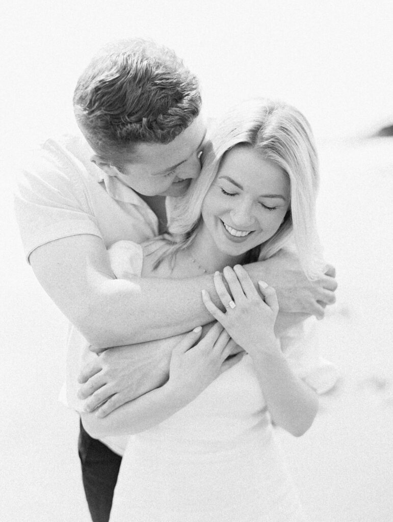 Malibu Engagement Session - A man embraces a smiling woman from behind. The woman shows off a ring on her finger, suggesting they are engaged or newly married. Both are happy and standing in a softly lit, bright setting. Copyright - Lisa Leanne Photography, California