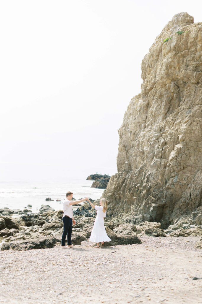 Malibu Engagement Session - A couple dances together on a rocky beach near a large cliff, with the ocean in the background. The woman wears a white dress and the man wears a white shirt and dark pants. The scene is bright and airy. Copyright - Lisa Leanne Photography, California
