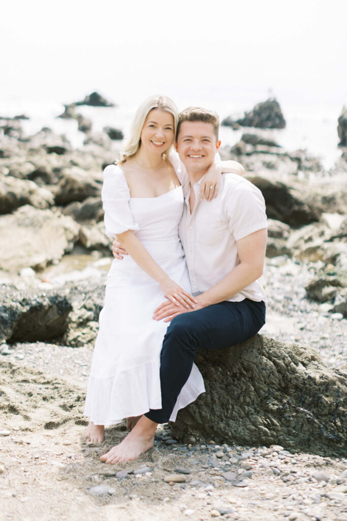 Malibu Engagement Session - A smiling couple sits on a rock at the beach. The woman wears a white dress and the man wears a light shirt and dark pants. Rocky shoreline and ocean waves appear in the background. Copyright - Lisa Leanne Photography, California