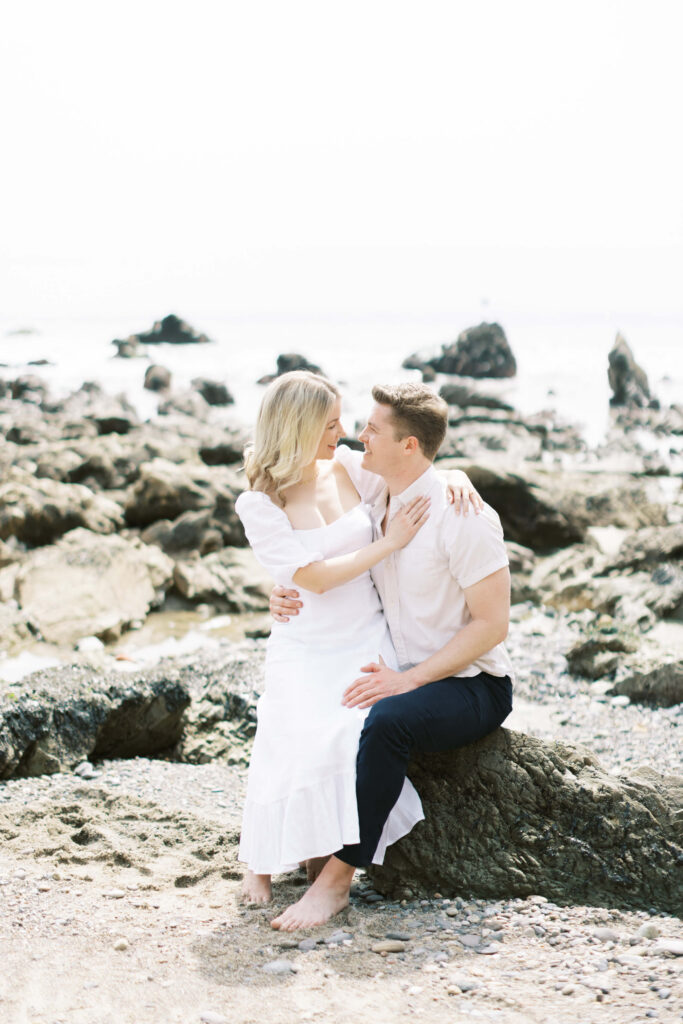 Malibu Engagement Session - A couple sits together on a rocky beach, embracing and smiling at each other. The woman wears a white dress, and the man wears a light shirt and dark pants. The rocky shore and calm water are visible in the background. Copyright - Lisa Leanne Photography, California