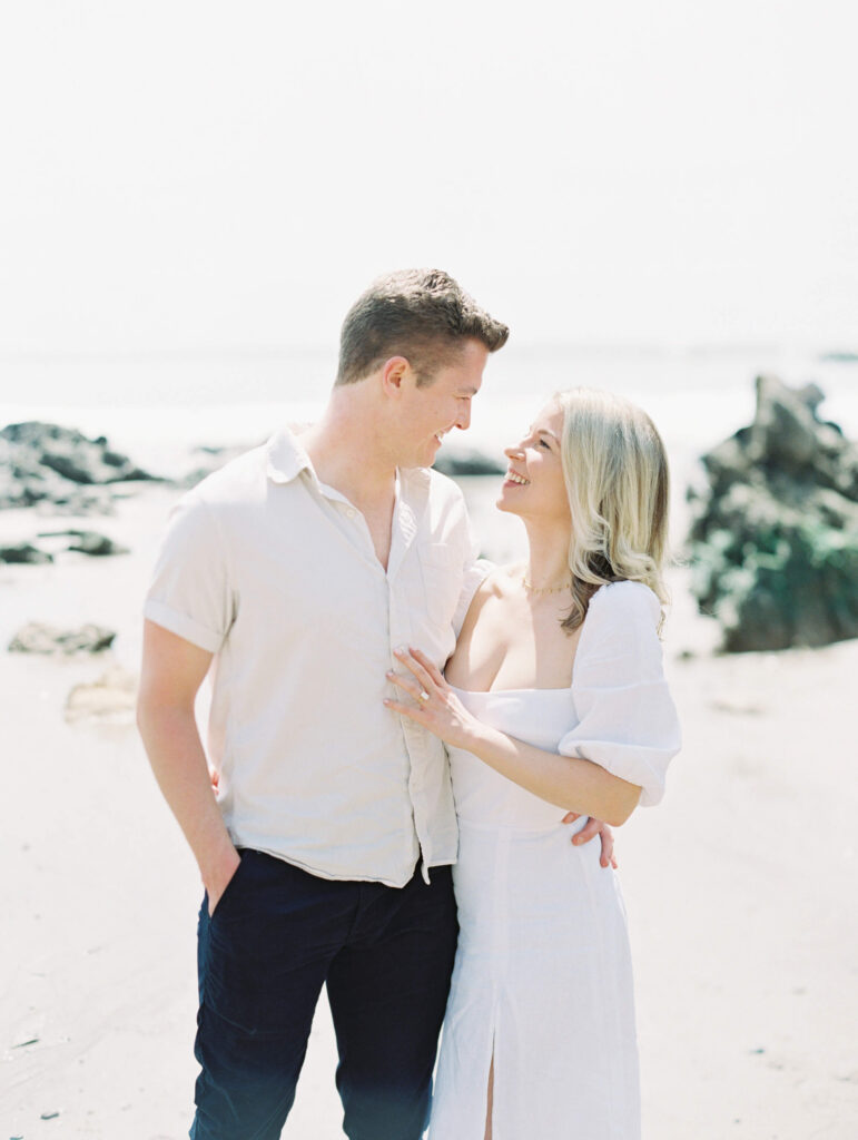 El Matador Beach Engagement Session - A smiling couple stands close together on a sandy beach, looking at each other. The woman wears a white dress and the man wears a light shirt and dark pants. Rocks and the ocean are visible in the background. Copyright - Lisa Leanne Photography, California