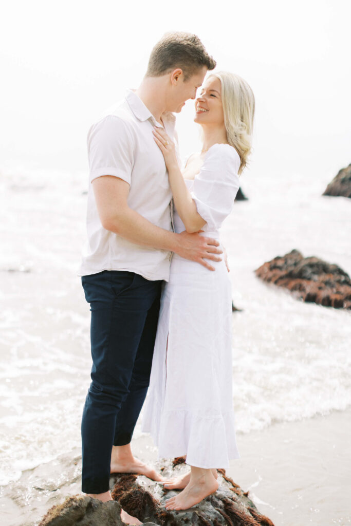 Malibu Engagement Session - A couple stands barefoot on rocks at the beach, embracing and smiling at each other. The woman wears a white dress and the man wears a light shirt and dark pants. Waves and rocks are visible in the background. Copyright - Lisa Leanne Photography, California