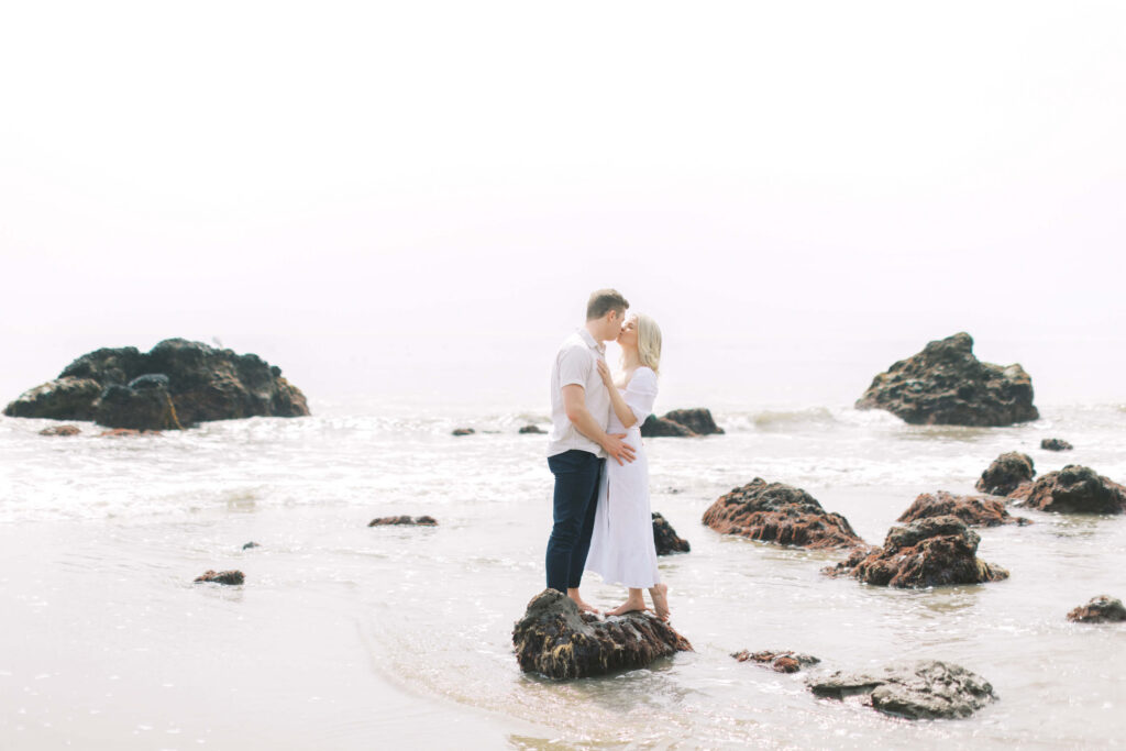 Malibu Engagement Session - A couple stands barefoot on rocks at the edge of the ocean, embracing and touching foreheads. The sky is bright and hazy, and dark rocks are scattered in the shallow water around them. Copyright - Lisa Leanne Photography, California