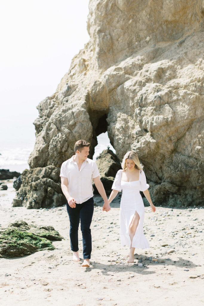 Malibu Beach Engagement Session - A couple walks hand in hand on a rocky beach, both wearing light-colored clothing. Behind them is a large rock formation with a small arch, and the ocean is visible in the background. Copyright - Lisa Leanne Photography, California