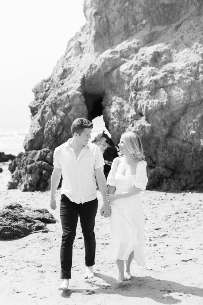 Malibu Beach Engagement Session - A couple walks barefoot on a sandy beach, holding hands and smiling at each other. They wear light-colored clothing, and a large rocky cliff is in the background. The image is in black and white. Copyright - Lisa Leanne Photography, California