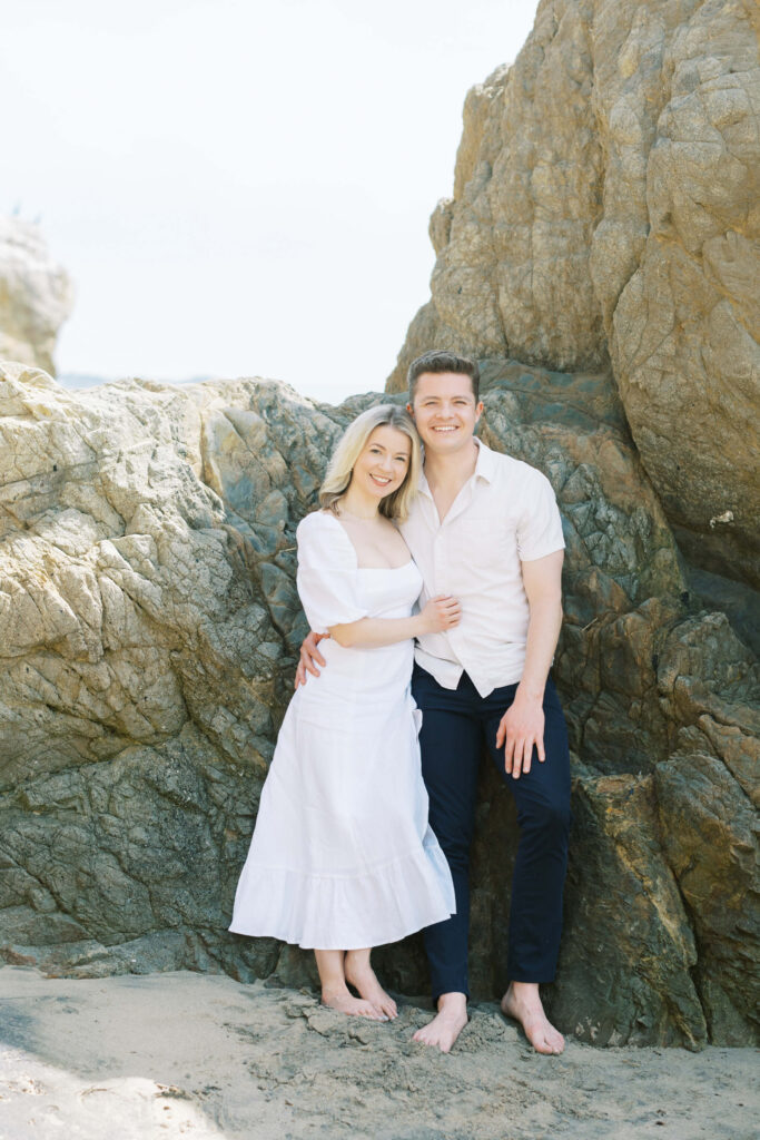 El Matador Beach Engagement Session - A smiling couple stands barefoot on a sandy beach, leaning against large rocks. The woman wears a white dress, and the man wears a light shirt with dark pants. They appear happy and relaxed in the natural, sunlit setting. Copyright - Lisa Leanne Photography, California