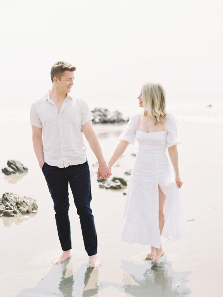 El Matador Beach Engagement Session - A couple holds hands and smiles at each other while walking barefoot on a sandy beach. The man wears a light shirt and dark pants, and the woman wears a white dress. Rocks and the ocean are visible in the background. Copyright - Lisa Leanne Photography, California