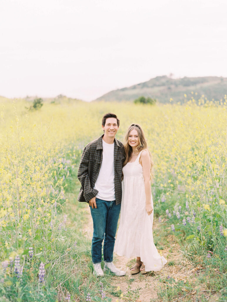 Malibu Engagement Session - Malibu Canyon - A young couple stands close together on a dirt path in a field of wildflowers, smiling at the camera. The man wears jeans, a T-shirt, and a plaid shirt, while the woman wears a flowing white dress. Hills are visible in the background. Copyright - Lisa Leanne Photography, California