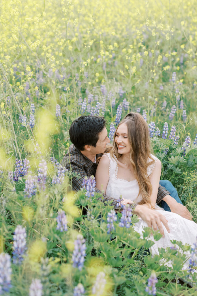 Malibu Engagement Session - Malibu Canyon - A couple sits closely together in a field of wildflowers, smiling and looking at each other. The woman wears a white dress, and the man wears a plaid shirt. Purple and yellow flowers surround them under soft natural light. Copyright - Lisa Leanne Photography, California