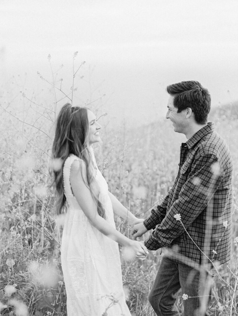 Malibu Engagement Session - Malibu Canyon - A couple stands in a field of tall wildflowers, holding hands and smiling at each other. The scene is in black and white, giving it a soft, romantic feel. Copyright - Lisa Leanne Photography, California