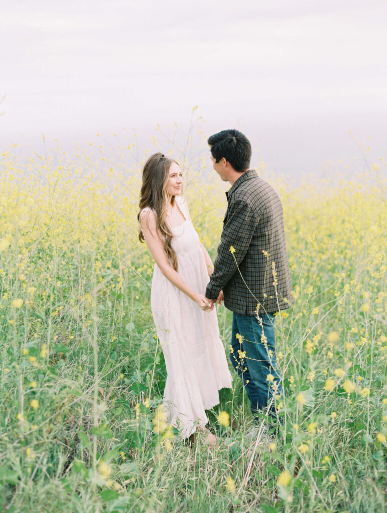 Malibu Engagement Session - Malibu Canyon - A couple stands in a field of yellow wildflowers, holding hands and facing each other. The woman wears a light, sleeveless dress and the man wears a plaid shirt and jeans. The scene is bright and serene. Copyright - Lisa Leanne Photography, California