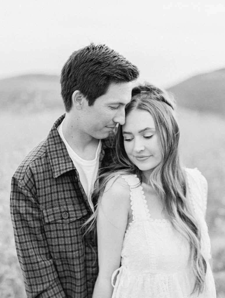 Malibu Engagement Session - Malibu Canyon - A black-and-white photo of a young couple standing in a field. The man, wearing a plaid shirt, stands behind the woman and leans his forehead gently against her temple. The woman, in a white dress, smiles softly with her eyes closed. Copyright - Lisa Leanne Photography, California