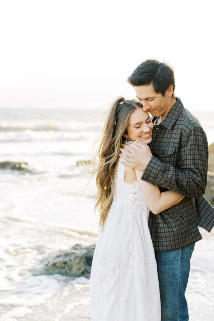 Malibu Engagement Session - El Matador Beach - A couple stands on the beach at sunset, embracing and smiling. The woman wears a white dress and the man wears a plaid shirt and jeans. Waves and rocks are visible in the background. Copyright - Lisa Leanne Photography, California