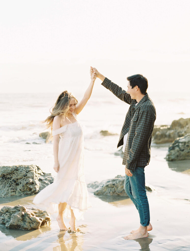 Malibu Engagement Session - El Matador Beach - A woman in a white dress and a man in jeans and a plaid shirt dance barefoot on a sandy beach near the water’s edge, surrounded by rocks, with soft sunlight illuminating the scene. Copyright - Lisa Leanne Photography, California