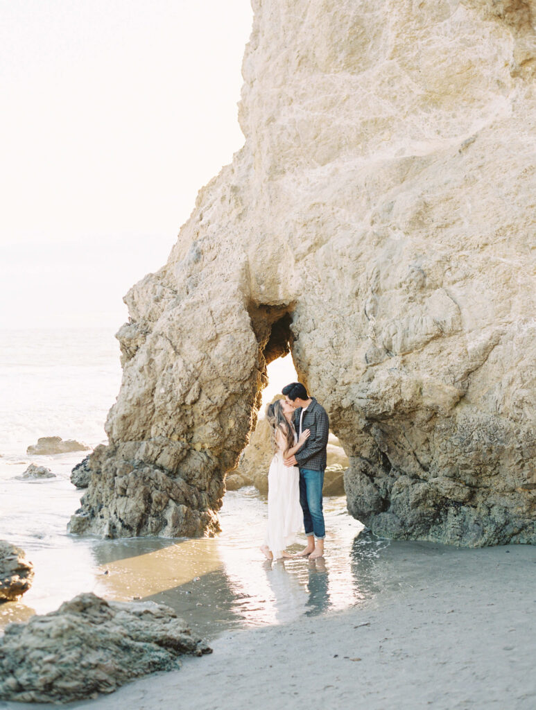 Malibu Engagement Session - El Matador Beach - A couple stands barefoot on the beach, embracing and touching foreheads in front of a large rocky archway at the edge of the ocean, with soft sunlight illuminating the scene. Copyright - Lisa Leanne Photography, California