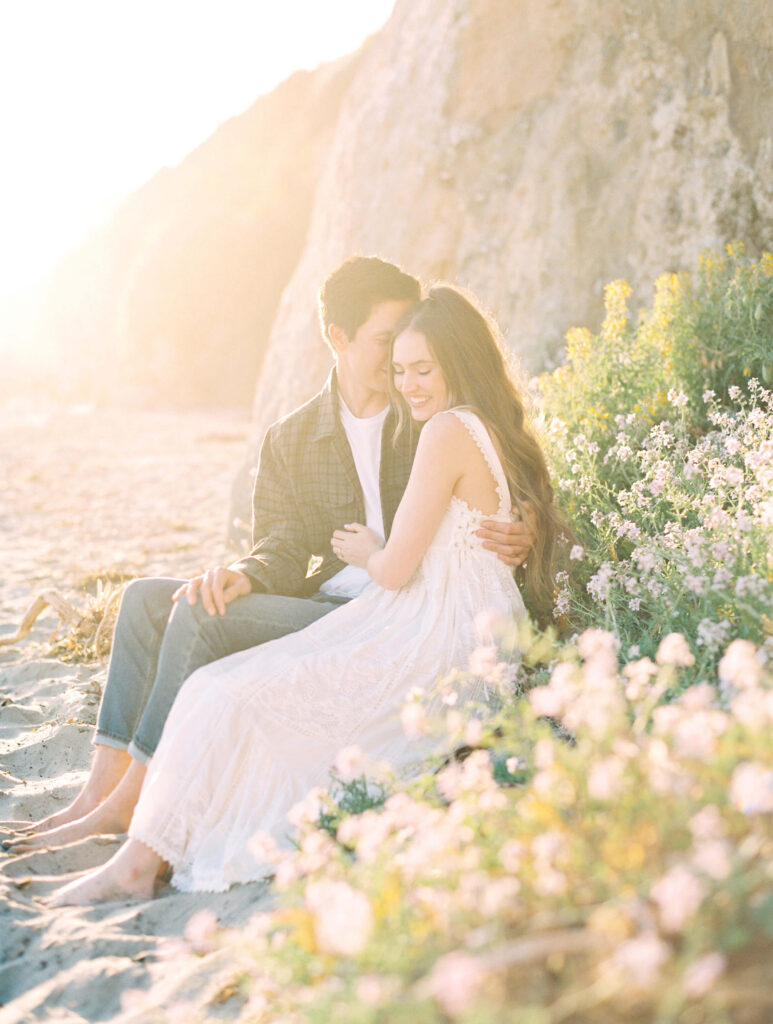 Malibu Engagement Session - El Matador Beach - A couple sits barefoot on a sandy beach near flowering bushes and a rocky cliff, embracing and smiling warmly at each other in soft, golden sunlight. Copyright - Lisa Leanne Photography, California