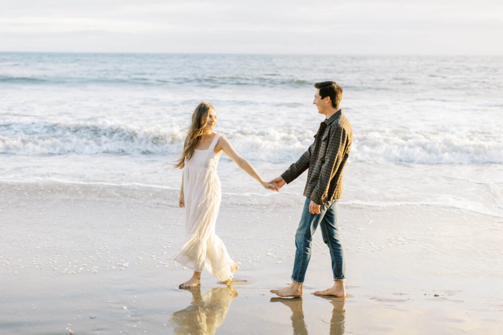 Malibu Engagement Session - El Matador Beach - A couple walks barefoot on the beach, holding hands and smiling at each other, with gentle waves and the ocean in the background. The woman wears a white dress and the man wears a plaid shirt and rolled-up jeans. Copyright - Lisa Leanne Photography, California