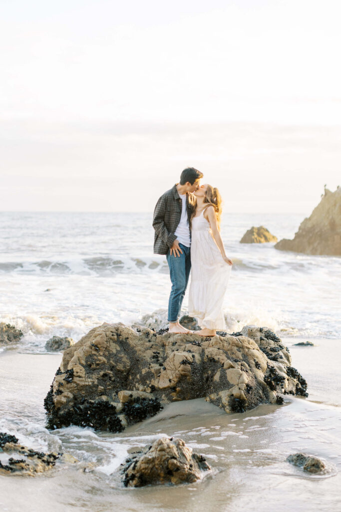 Malibu Engagement Session - El Matador Beach - A couple stands barefoot on a rock at the beach, facing each other and about to kiss. The ocean waves and distant rocks are visible behind them under a soft, cloudy sky. Copyright - Lisa Leanne Photography, California