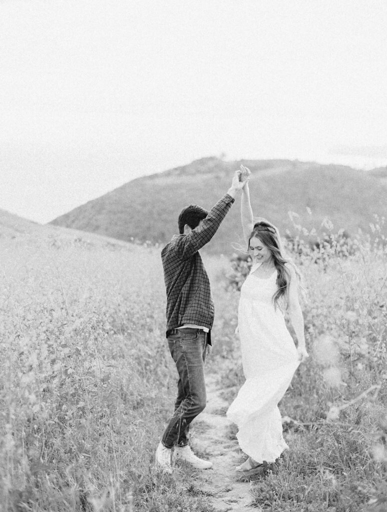 Malibu Engagement Session - Malibu Canyon - A couple dances on a narrow path in a field of tall grass, with rolling hills in the background. The person on the left wears a plaid shirt and jeans, while the person on the right wears a flowing dress. The image is in black and white. Copyright - Lisa Leanne Photography, California
