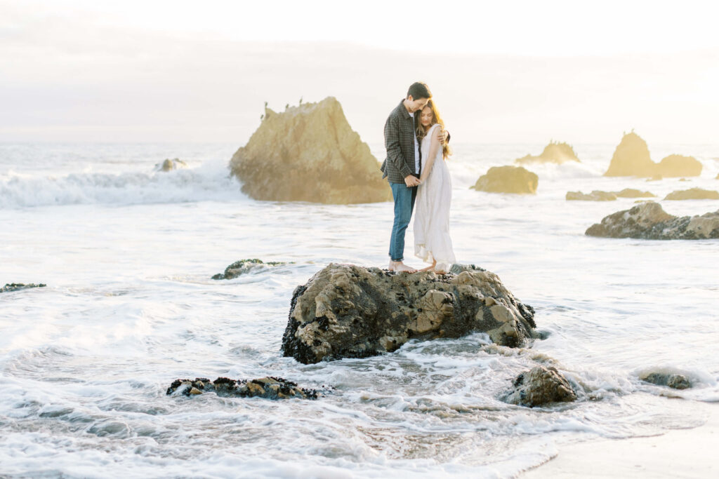 Malibu Engagement Session - El Matador Beach - couple stands barefoot on a rock surrounded by ocean waves, embracing and touching foreheads, with large rocks and gentle sunlight in the background. Copyright - Lisa Leanne Photography, California