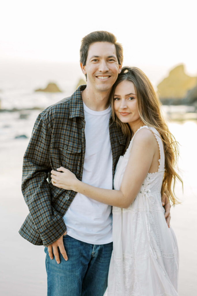 Malibu Engagement Session - El Matador Beach - A young couple stands close together on a beach, smiling at the camera. The man wears a plaid shirt and jeans, while the woman wears a white dress. The ocean and large rocks are visible in the background. Copyright - Lisa Leanne Photography, California