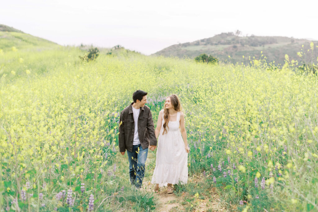 Malibu Engagement Session - Malibu Canyon - A couple walks hand in hand along a narrow dirt path through a field of tall yellow wildflowers, with green hills and a cloudy sky in the background. Copyright - Lisa Leanne Photography, California