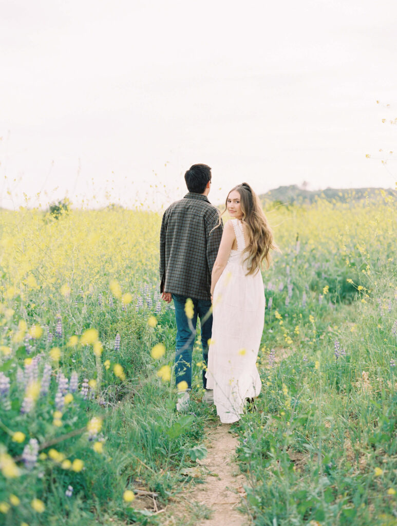 Malibu Engagement Session - Malibu Canyon - A couple walks along a narrow path through a field of tall wildflowers. The woman in a long white dress looks back over her shoulder, while the man in a plaid shirt faces forward. The scene is bright and serene. Copyright - Lisa Leanne Photography, California