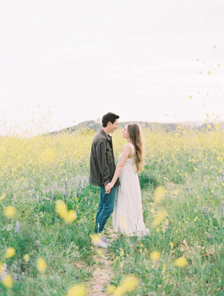 Malibu Engagement Session - Malibu Canyon - A couple stands facing each other, holding hands in a field of yellow wildflowers. The woman wears a long white dress, and the man wears a plaid shirt and jeans. The scene is bright and peaceful, with flowers in the foreground. Copyright - Lisa Leanne Photography, California