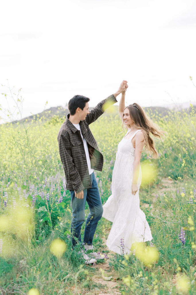 Malibu Engagement Session - Malibu Canyon - A couple stands in a field of wildflowers; the man twirls the woman, who wears a white dress, while both smile and enjoy the sunny, natural setting. Copyright - Lisa Leanne Photography, California