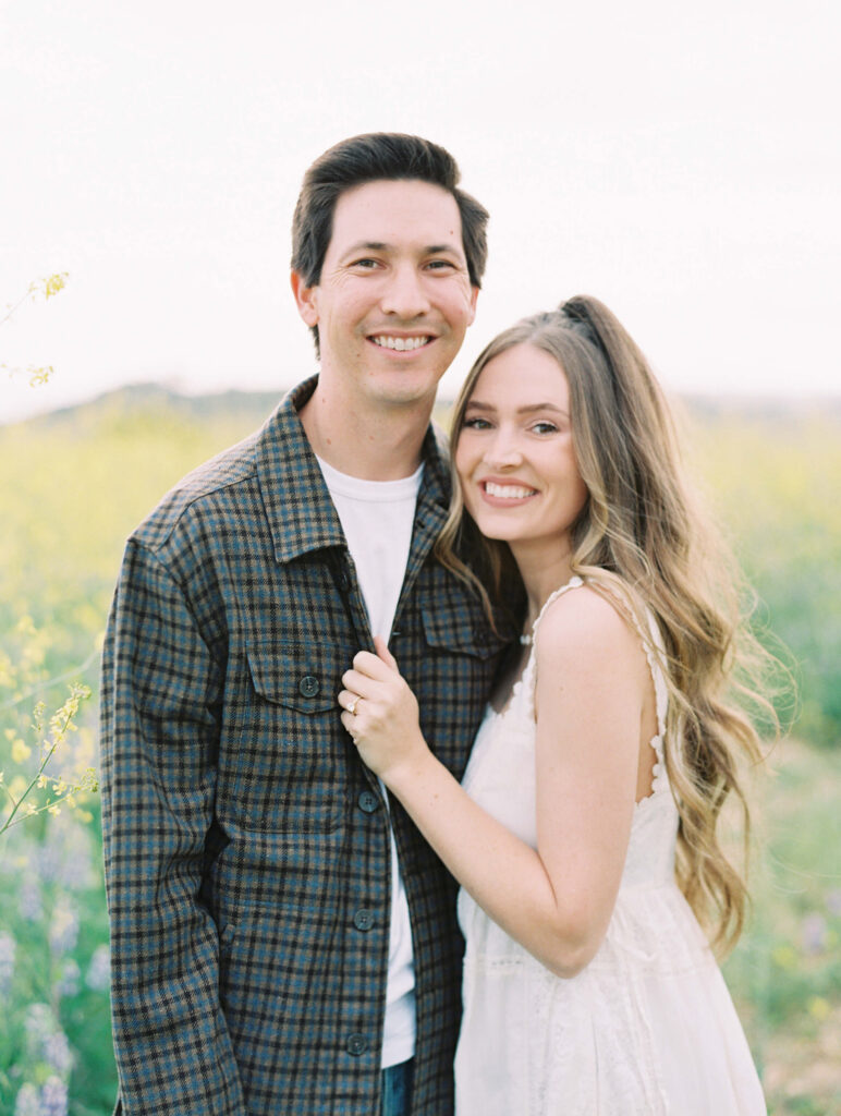 Malibu Engagement Session - Malibu Canyon - A young couple stands close together outdoors in a field of yellow flowers, smiling at the camera. The man wears a plaid jacket and the woman wears a white dress with long, wavy hair. The scene is bright and cheerful. Copyright - Lisa Leanne Photography, California